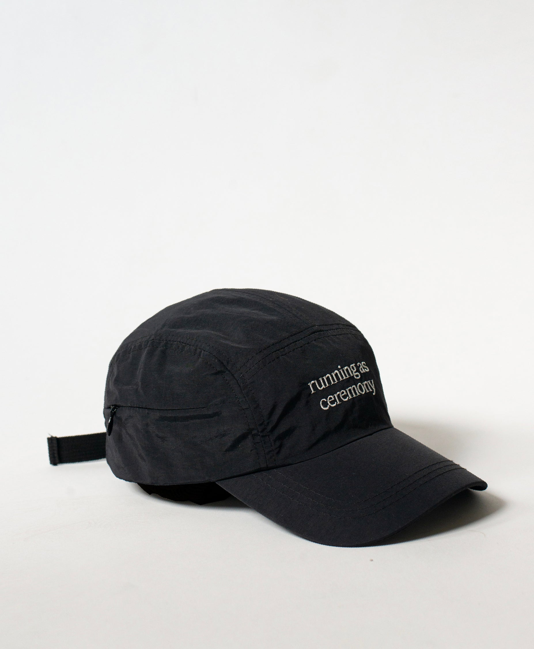 Three-quarter view of the Philos Running Cap in black, with text in white that reads "running as ceremony" and a discreet key pocket with a zipper.