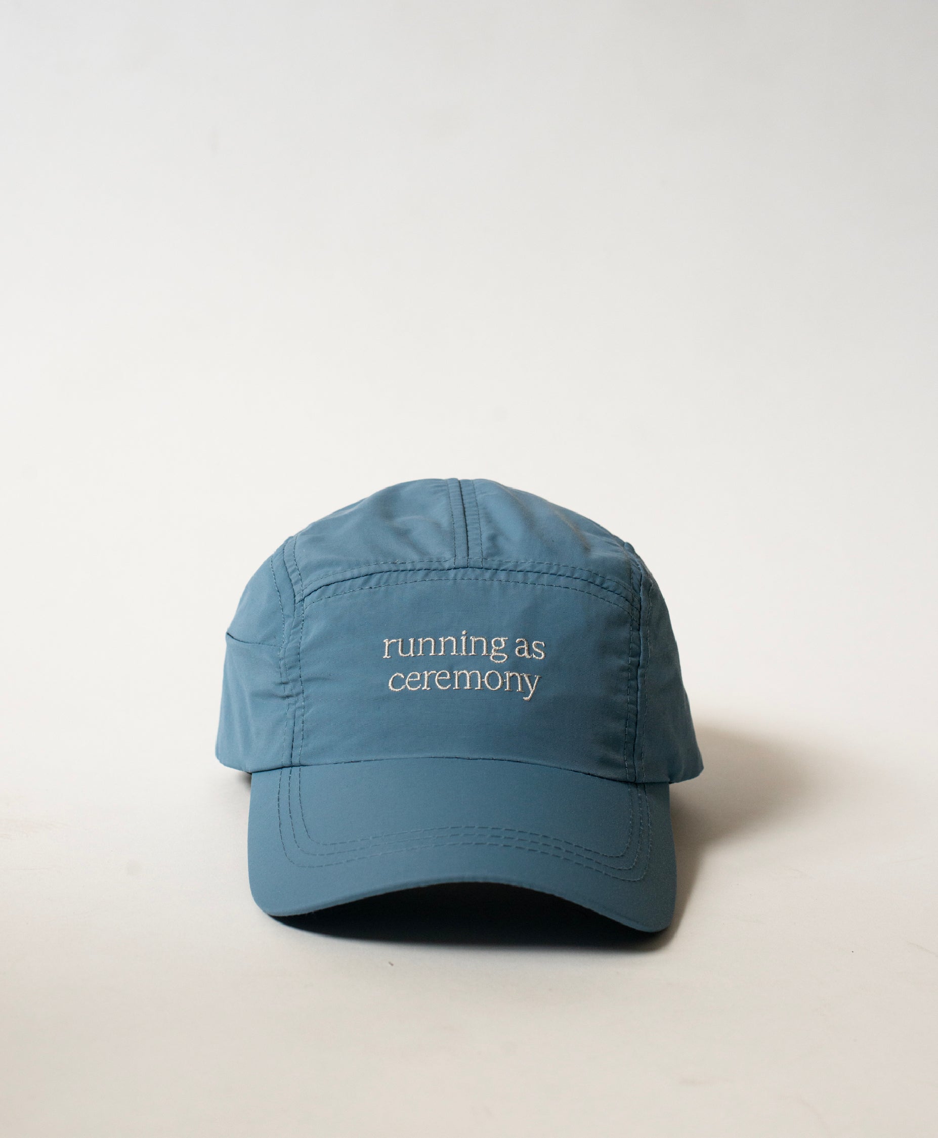 Front view of the Philos Running Cap in sky blue, made from water-resistant fabric. Features white text "running as ceremony" and a discreet key pocket.