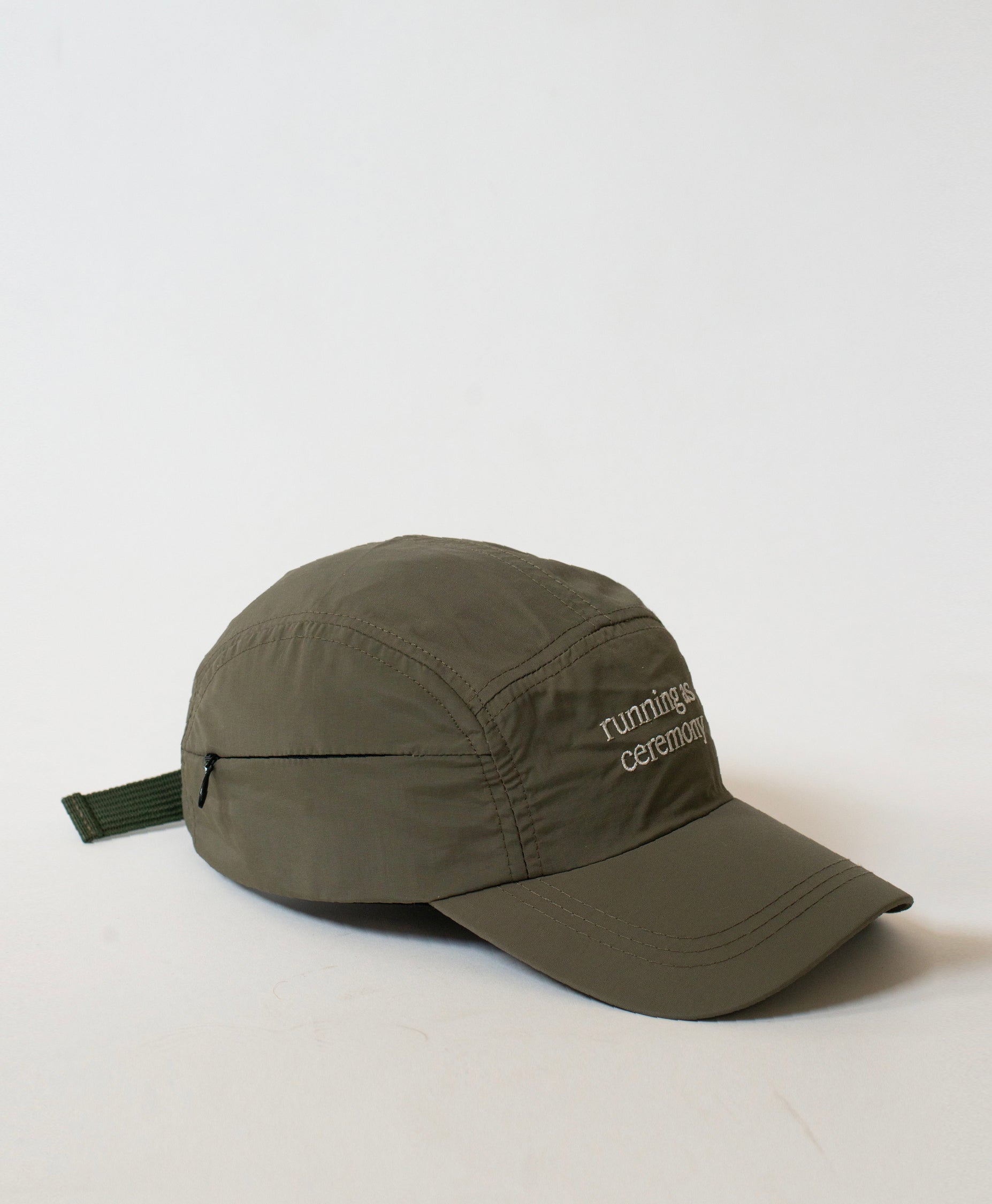 Three-quarter view of the Philos Running Cap in olive green, with text in white that reads "running as ceremony" and a discreet key pocket with a zipper.