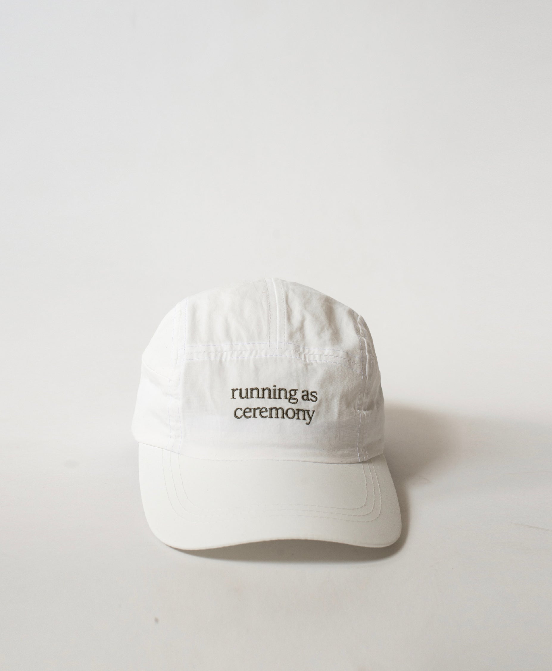 Front view of the Philos Running Cap in white, made from water-resistant fabric. Features gray text "running as ceremony" and a discreet key pocket.