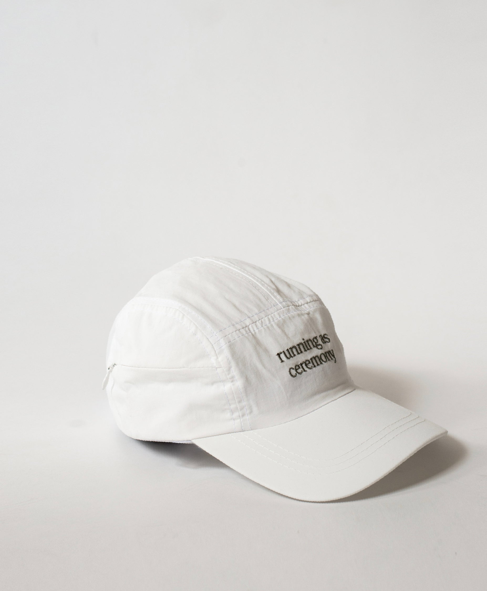 Three-quarter view of the Philos Running Cap in white, with text in white that reads "running as ceremony" and a discreet key pocket with a zipper.