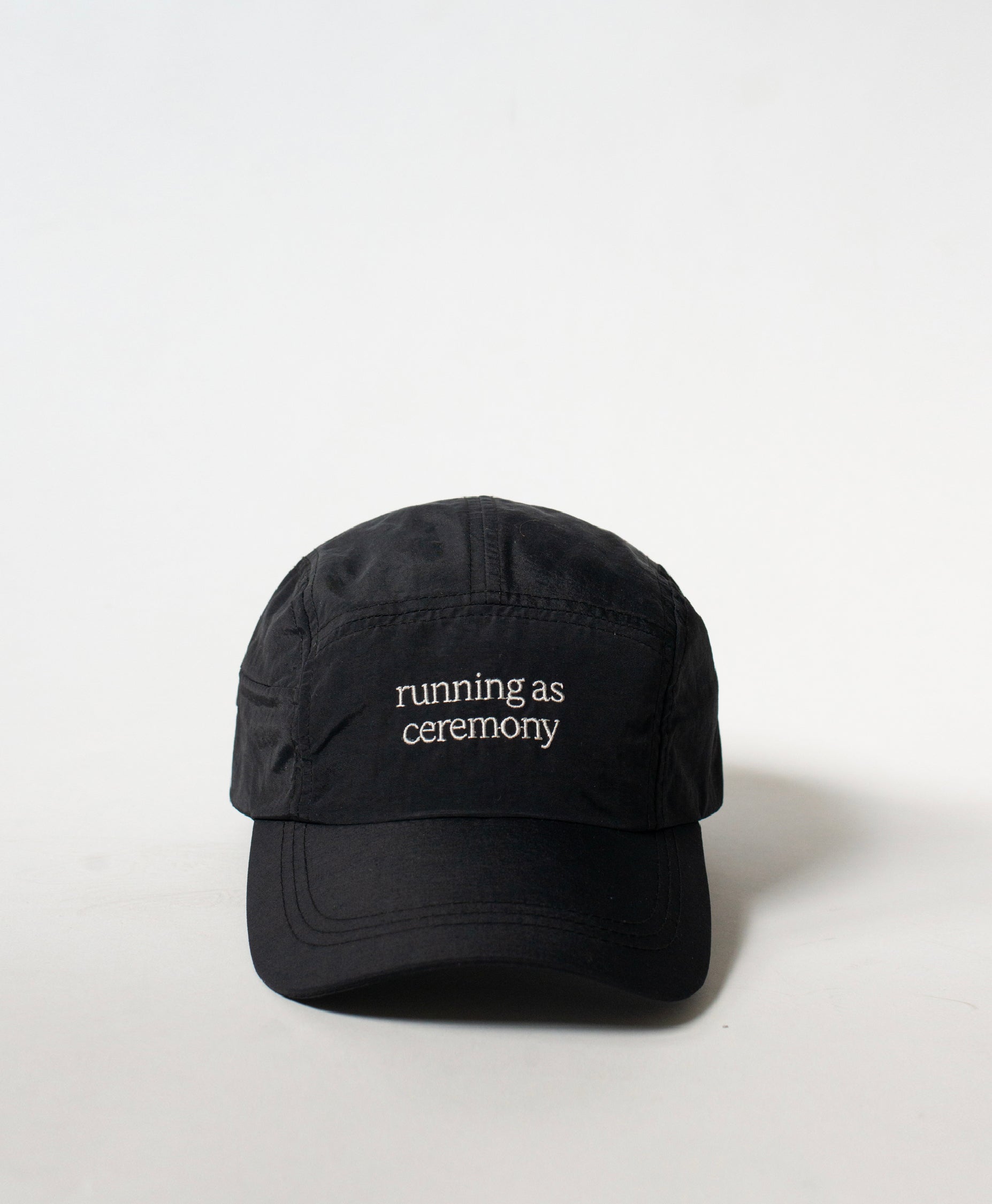 Front view of the Philos Running Cap in black, made from water-resistant fabric. Features white text "running as ceremony" and a discreet key pocket.