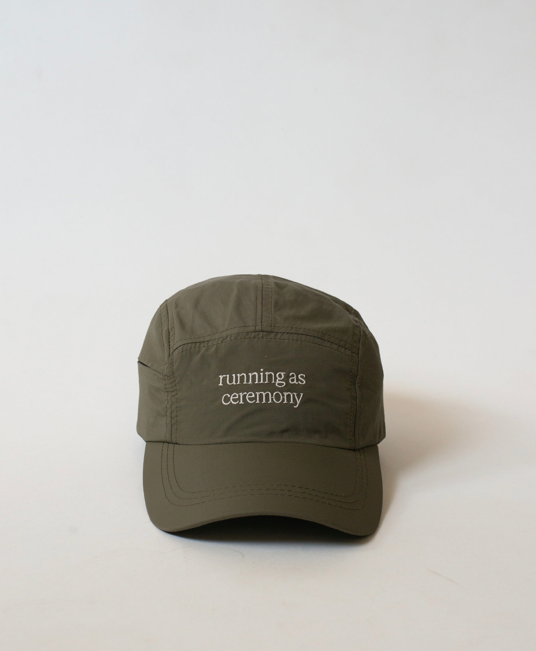 Front view of the Philos Running Cap in olive green, made from water-resistant fabric. Features white text "running as ceremony" and a discreet key pocket.